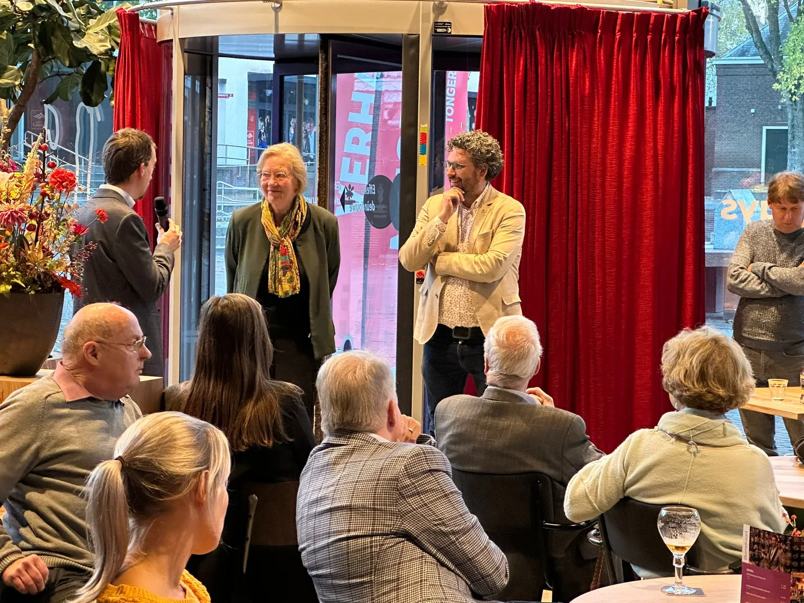 Arwen van Gestel begeleidt een bijeenkomst voor publiek in een theaterachtige setting met rode gordijnen